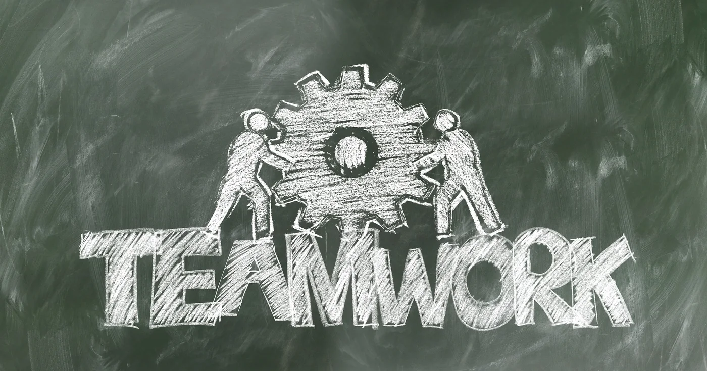 Tafelzeichnung: zwei Personen bewegen gemeinsam ein Zahnrad, darunter der Schriftzug „Teamwork“